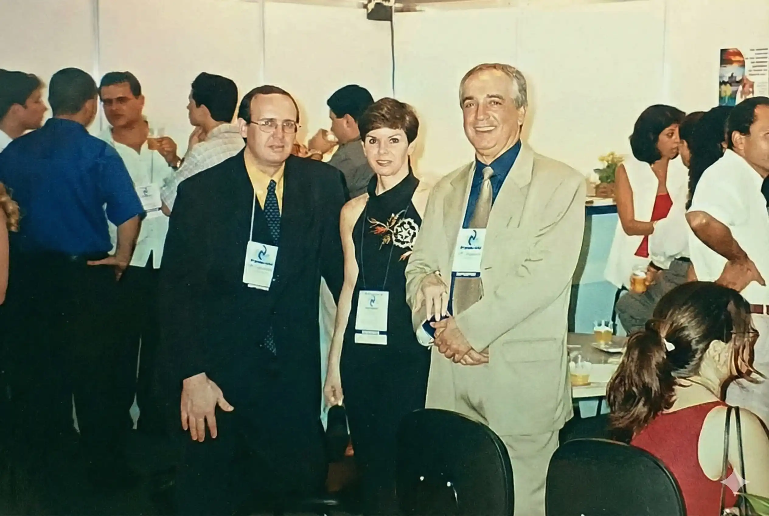 Três pessoas com crachás de evento posando para foto em um encontro fechado, com outros participantes ao fundo