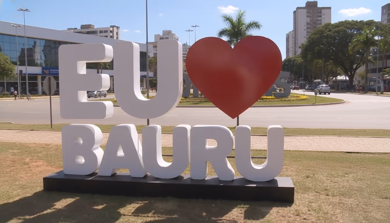 Escultura ao ar livre com as letras brancas EU e BAURU e um coração vermelho no lugar do verbo amar, sobre um gramado com prédios e árvores ao fundo
