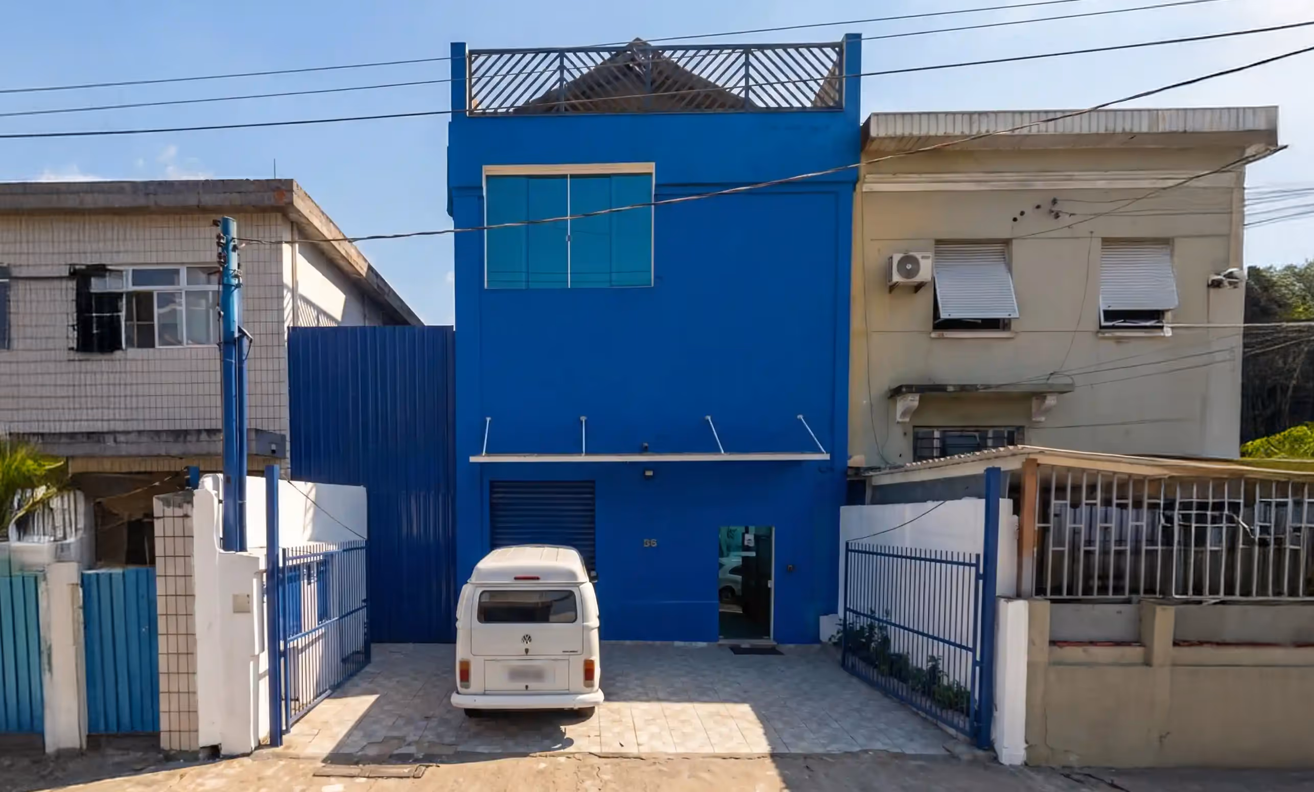 Van branca estacionada em frente a um prédio azul de dois andares com portão, em um dia de sol
