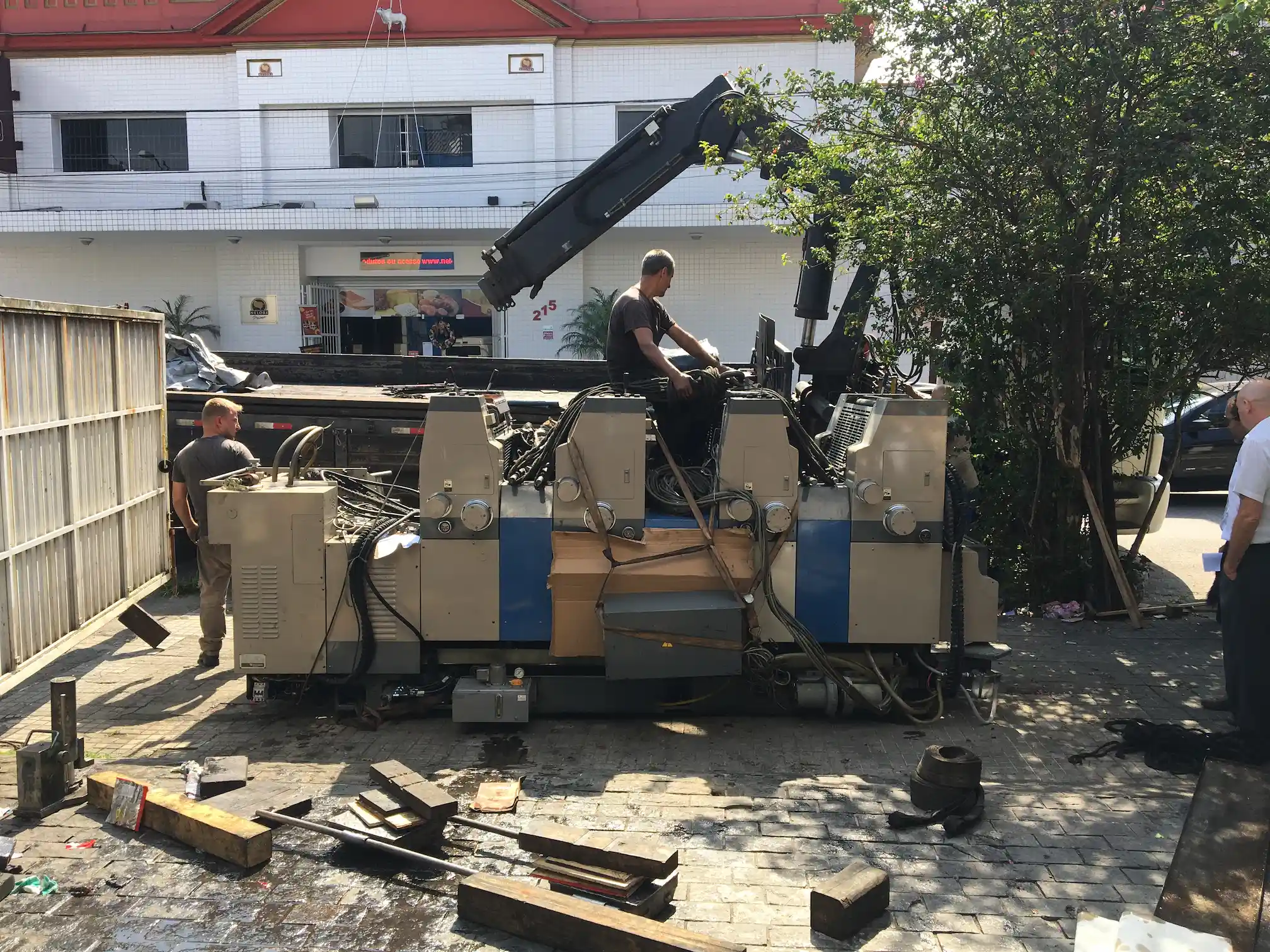 Três homens movendo ao ar livre uma grande máquina industrial com um guindaste acoplado a um caminhão prancha